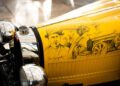 A yellow vintage car hood features sketches of two men in hats, a classic car, and signatures of "Jean Bugatti" and "Ettore Bugatti," capturing the spirit of Hedley Studios and the timeless elegance of the Bugatti Baby II.