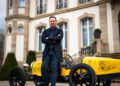 A man in a black suit stands confidently with arms crossed in front of a yellow vintage-style car, reminiscent of the Bugatti Baby II, outside a large, elegant building. A seamless collaboration with Hedley Studios brings an air of sophistication to the scene.