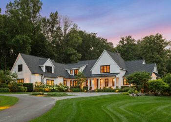 A large, modern farmhouse-style home with white siding and dark trims is surrounded by trees and a manicured lawn at dusk. A curved driveway leads to the entrance.