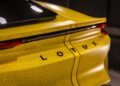 Rear view of a yellow Lotus Emira, showcasing a distinctive text pattern design. The brand name "LOTUS" is prominently visible on the back, hinting at its turbocharged performance.