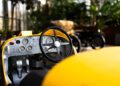 In a detailed close-up of a Bugatti Baby II's interior, the yellow car reveals its steering wheel and dashboard with precise gauges. The blurred background, captured by Hedley Studios, subtly showcases plants and another vehicle.