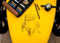 A sketch of a Bugatti Baby II rests on a yellow surface, framed by colored pencils and markers. Nearby, a black and yellow Hedley Studios coffee cup completes the artistic arrangement.