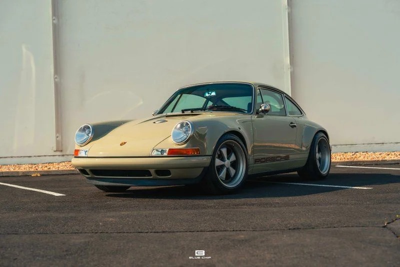A classic air-cooled 911 Porsche sports car in beige is parked on an asphalt surface in front of a plain white wall.