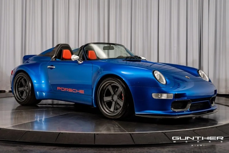 A blue Porsche Speedster inspired by Singer, with black wheels and a red-accented interior, is displayed on a rotating platform with a gray curtain backdrop.