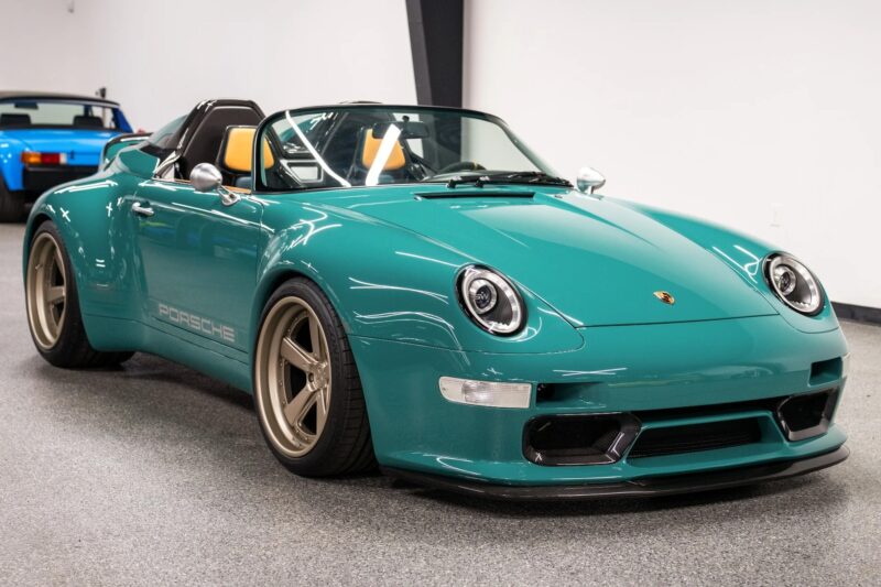 A teal air-cooled 911 Porsche convertible with tan seats and bronze wheels is parked in a clean indoor showroom, with a blue car visible in the background.