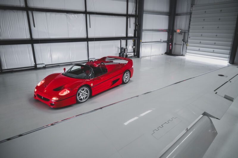 A Ferrari F50, gleaming in its iconic red, is parked inside a large industrial garage with white walls and a closed roller door, awaiting the Miami Auction at RM Sotheby's.