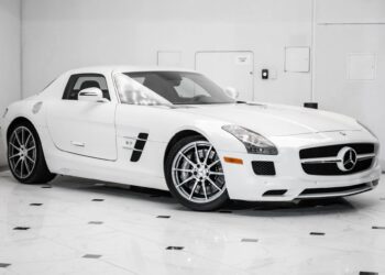 A white Mercedes-Benz SLS AMG luxury car is parked indoors on a shiny, tiled floor with a plain white wall in the background.
