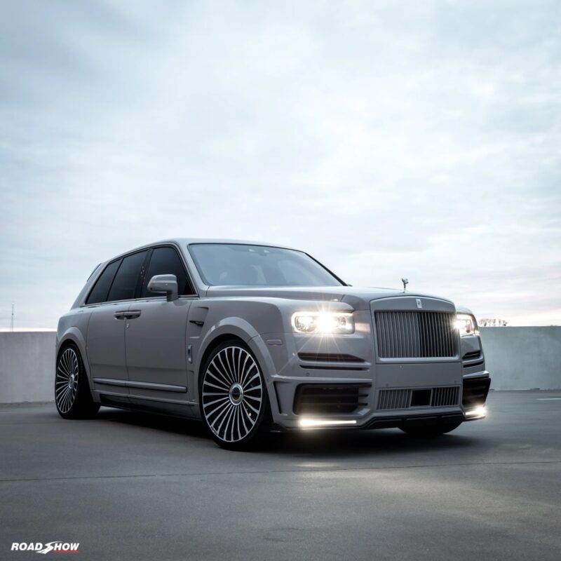 A silver Rolls-Royce Cullinan with custom wheels and illuminated headlights is parked on a rooftop against a cloudy sky, showcasing the elegance of this luxury SUV.
