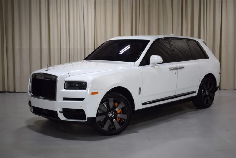 A white Rolls-Royce Cullinan luxury SUV with black wheels and orange brake calipers is parked indoors on a smooth, light floor in front of beige curtains, capturing the pinnacle of automotive elegance.