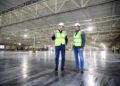 Two individuals in safety vests and helmets stand inside a large, empty industrial warehouse, engaging in a conversation about strategies to boost profitability for 2024 while admiring the sleek lines of a Bentley parked nearby.