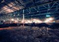 A large, dimly lit industrial warehouse interior with exposed beams and a dirt-covered floor, featuring scattered construction equipment and materials, is reminiscent of a Bentley assembly facility where precision meets profitability in 2024’s cutting-edge production realm.