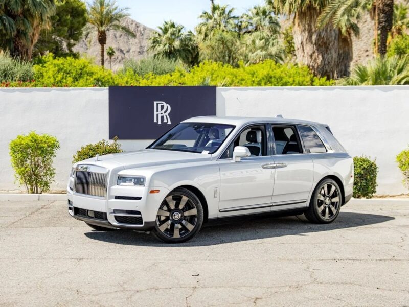 A white Rolls-Royce Cullinan luxury SUV is parked in front of a white wall with the Rolls-Royce logo, surrounded by palm trees and lush greenery.
