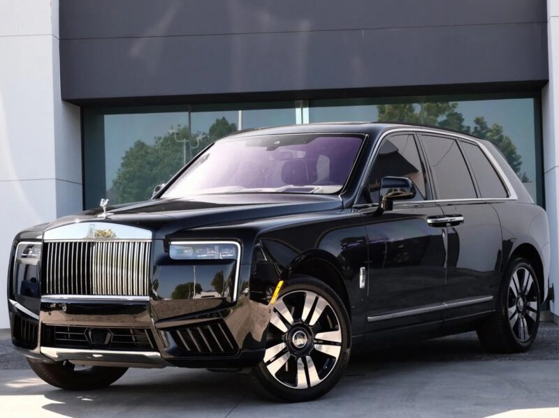 A black Rolls-Royce Cullinan, the pinnacle of luxury SUV design, is parked in front of a modern building with large glass windows and a gray facade.