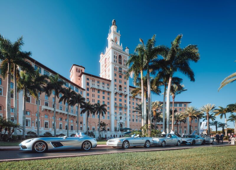 A row of sleek Silver Stars parked in front of a grand hotel with a towering structure, surrounded by swaying palm trees under a clear blue sky.