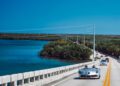 A sleek Mercedes-Benz convertible glides across a long bridge over blue waters, surrounded by lush greenery. Silver stars of ModaMiami 2025 shine as other cars form a picturesque backdrop.