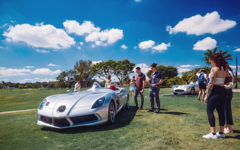 People gathered around a sleek Mercedes-Benz on a grassy field under a blue sky with clouds, the silver star emblem gleaming in the sun—a scene capturing the spirit of innovation anticipated at ModaMiami 2025.