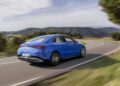 A blue Mercedes-Benz New CLA cruises on a winding road through a hilly landscape, surrounded by trees under a partly cloudy sky, showcasing its groundbreaking tech.