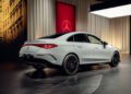 A sleek white Mercedes-Benz New CLA is parked indoors, showcasing a luxurious ambiance. With a cityscape backdrop and branded logo on the wall, it embodies groundbreaking tech and elegance from every side view angle.