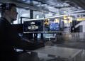 A man in a dimly lit room observes dual computer monitors displaying digital content, viewed through glass, reminiscent of the precision found in a Mercedes-Benz.