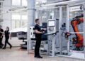 A person operates a control panel near a robotic arm in a modern industrial setting, surrounded by the precision engineering of Mercedes-Benz. In the background, two people walk past, highlighting the seamless integration of AI technology in this advanced environment.