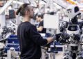 In a bustling factory, a man engages with a state-of-the-art humanoid robot. Surrounding machines and workers hum in the background, embodying the precision and innovation reminiscent of Mercedes-Benz's cutting-edge technologies.