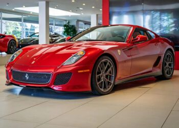 A red Ferrari 599 GTO, a must-have for any supercar collection, is displayed in a showroom with large windows and other cars in the background.