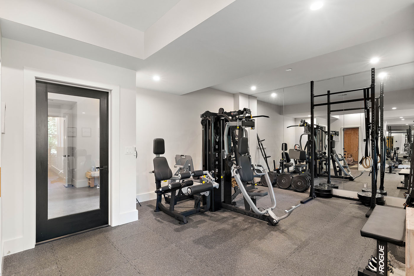 Gym rumah modern dengan mesin berat, rak dumbel, dan cermin besar. Kamar memiliki dinding putih, lantai bertekstur, dan pencahayaan yang cerah. Sebuah pintu terlihat di sebelah kiri.