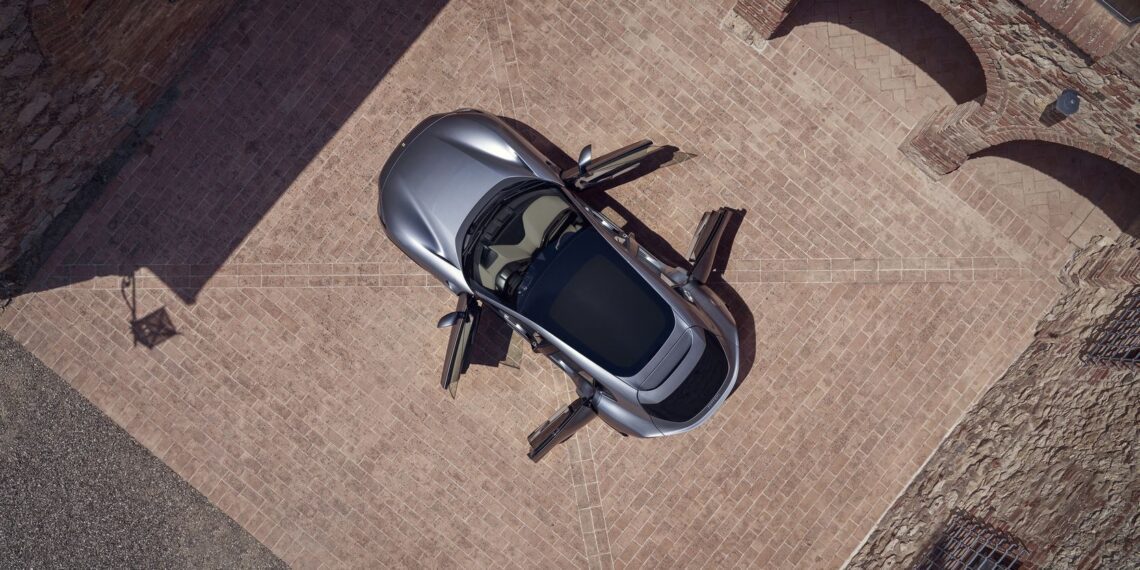 Overhead view of a sleek silver Ferrari sports car with its doors open, parked on a brick driveway surrounded by brick buildings.