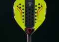 A yellow and black carbon padel racquet with red accents, showcasing logos in the center, stands upright on a white surface against a black background, reminiscent of Lamborghini's sleek design.