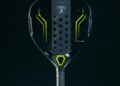 A black and yellow pickleball paddle, akin to a Lamborghini in sleekness, features a textured grip and perforations on the face. It stands upright on a white circular platform against a dark background, exuding luxury akin to a Babolat carbon padel racquet.
