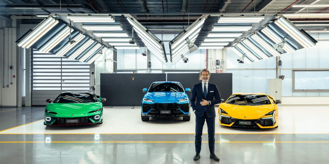 A man in a suit stands in front of three colorful sports cars—green, blue, and yellow Lamborghinis—inside a well-lit showroom with industrial ceiling lights, showcasing their anticipated 2024 sales.