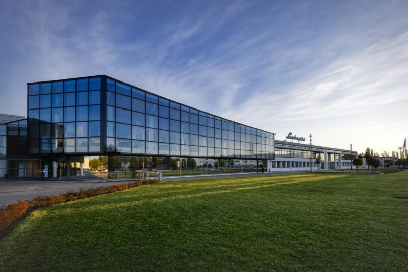 A sleek, modern glass-fronted industrial building with a Lamborghini parked out front stands proudly, its sign gleaming under a clear sky. Surrounded by a manicured lawn, it’s the perfect setting for efficient deliveries or high-powered sales meetings.