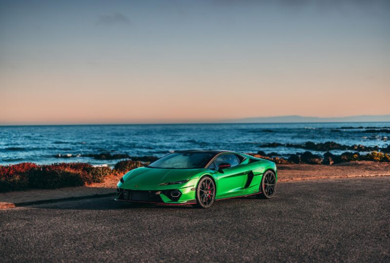 A green Lamborghini sportscar is parked on a coastal road, with the ocean and horizon offering a breathtaking backdrop under the clear sky.