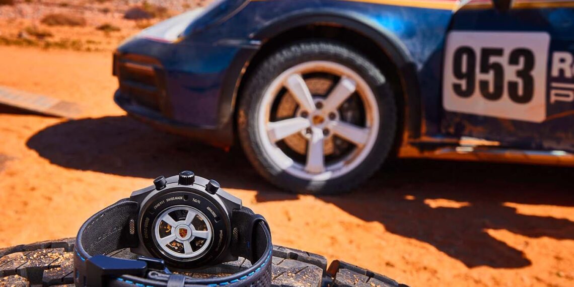 A sleek wristwatch with a black band rests on a tire in the desert, set against the backdrop of a race car labeled "953," capturing the essence of Porsche Design.