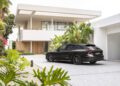 A modern house with large windows and a spacious driveway featuring a sleek black Audi sports car, surrounded by tropical plants and greenery.