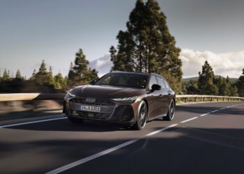 A sleek, dark-colored Audi A6 Avant Wagon glides effortlessly along a forested road under a partly cloudy sky, its hybrid engine whispering with every turn.