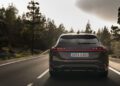 Rear view of a sleek black Audi A6 Avant Wagon gliding effortlessly down a forested road, surrounded by trees on both sides and a cloudy sky painting the backdrop.