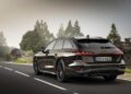 A black Audi A6 Avant cruises down a scenic road, surrounded by trees and rocks under a cloudy sky.