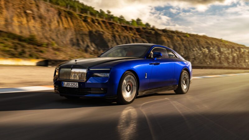 A blue and black luxury car driving on a road with a rocky hillside and trees in the background.