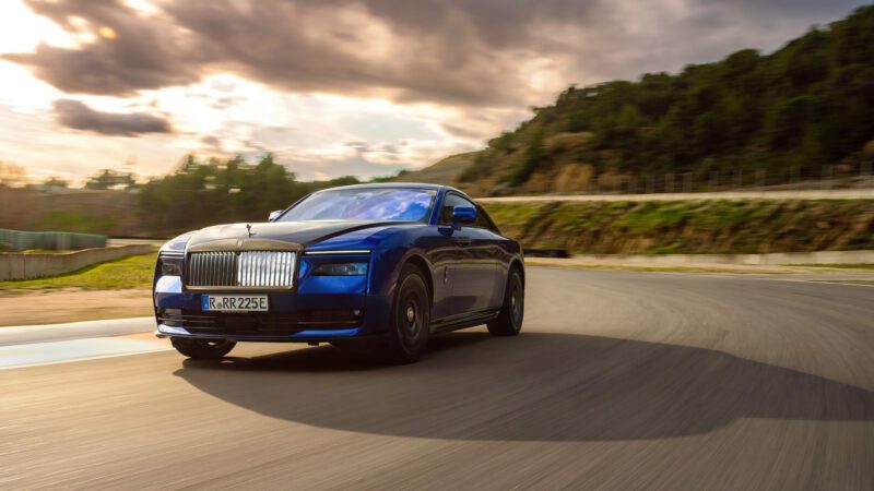 Blue luxury car driving on a curvy road with trees and a cloudy sky in the background.