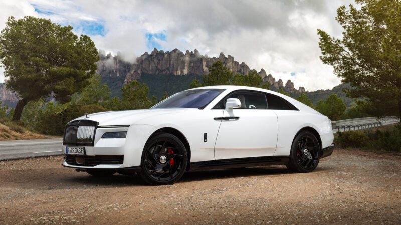 A white luxury car, rumored to be the Black Badge Spectre, is elegantly parked on a gravel area with trees and rocky hills in the background under a veiled cloudy sky.