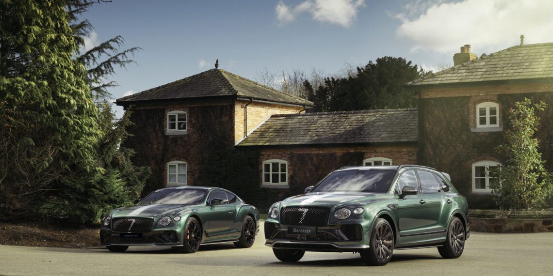 Two green luxury cars from the Le Mans Collection are parked on a driveway in front of a rustic house, surrounded by trees under a partly cloudy sky.