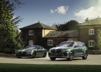 Two green luxury cars from the Le Mans Collection are parked on a driveway in front of a rustic house, surrounded by trees under a partly cloudy sky.