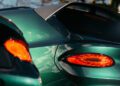 Close-up of a green sports car's rear, highlighting the taillight and a carbon fiber spoiler, reminiscent of the Le Mans racing spirit. Reflections of the sky dance across its sleek surface.