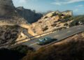 An Aston Martin Vanquish speeds along a scenic mountain road, navigating rocky terrain with ease. Under a partly cloudy sky, this fastest convertible glides past distant peaks, embodying elegance and power as only a front-engined convertible can.