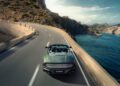 An Aston Martin Vanquish, the fastest front-engined convertible, glides along a coastal winding road with cliffs on one side and the ocean on the other under a clear sky.