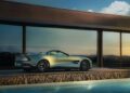 An Aston Martin Vanquish, the front-engined convertible, is parked beside a modern glass-fronted building, reflecting the sunset sky.