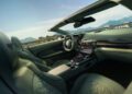 The interior of this front-engined convertible, an Aston Martin Vanquish, boasts green leather seats and a sleek modern dashboard, parked with a scenic mountain view in the background.
