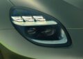 Close-up of a sleek Aston Martin Vanquish headlight with a modern, angular design and multiple LED elements. The car body, one of the fastest convertibles, boasts a smooth, metallic green finish.