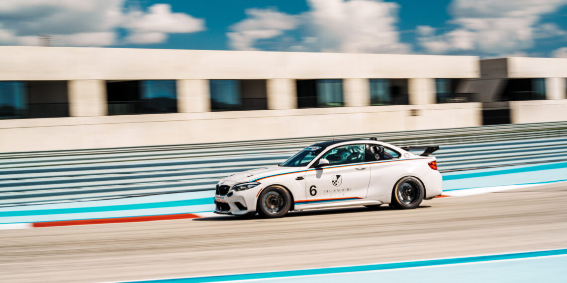 A sleek white race car races around the racetrack at Concours Club, its engine roaring in front of a modern building under a blue sky with scattered clouds.